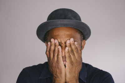 Man wearing hat covering face with hands against gray background