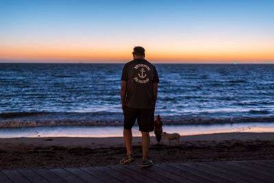 Rear view of man standing on beach against sky during sunset