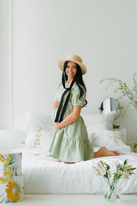 Pregnant girl dressed in a green dress and hat is sitting on the bed and holding her tummy