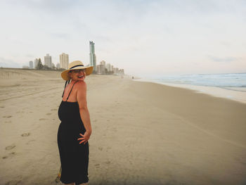 Happy woman wearing black dress standing at beach by city against sky