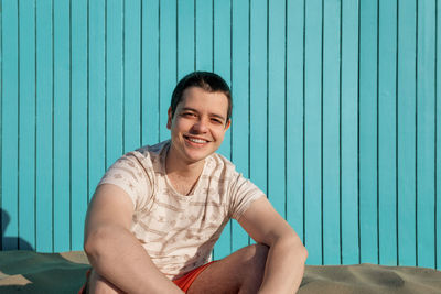 Young man sitting in sand beach smiling at camera in a turquoise wall