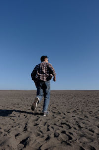 Full length rear view of man running on sand at desert against clear sky during sunny day