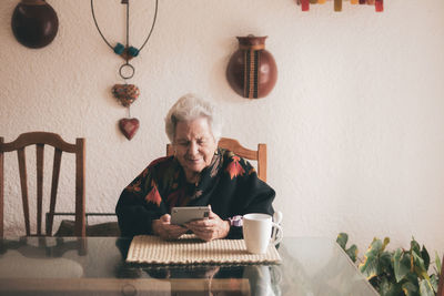 Smiling elderly female wearing warm clothes sitting at table with tablet and cup of tea looking at screen