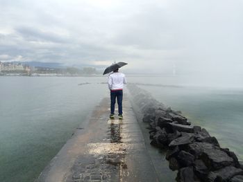 Full length rear view of man with umbrella walking on pier amidst sea during rainy season