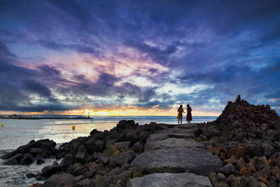 Men on rock at beach against sky during sunset