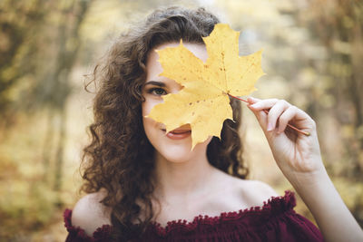 Beautiful girl with curly dark hair in a maroon top in a park holding maple leaf in front of face