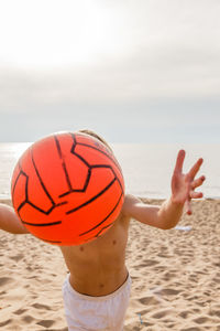 Boy throwing orange ball at beach against sky