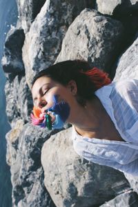 Woman with closed eyes holding tassel in mouth while sitting on rock at beach