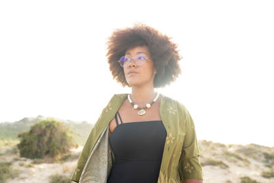 Dreamy young african american female with curly hair wearing stylish summer clothes with necklace and eyeglasses looking away while standing in sunlight on seashore in summertime