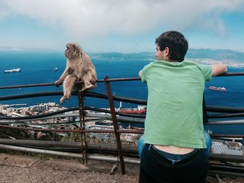 Rear view of man by monkey sitting on railing against harbor