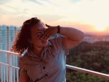 Portrait of young woman standing by railing against sky during sunset