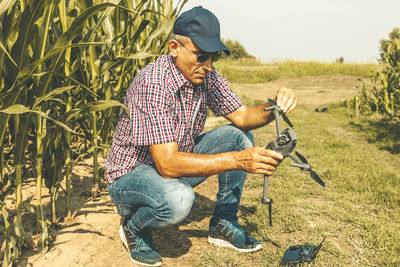 Full length of mature man wearing sunglasses holding drone while crouching on field against sky