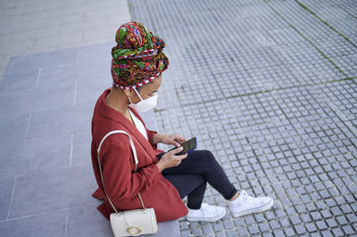 Woman wearing protective face mask and headscarf using mobile phone while sitting on footpath