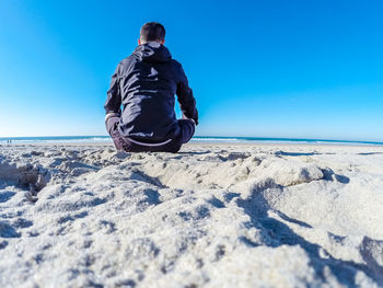 Rear view of man sitting on beach against clear blue sky