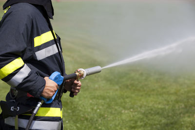 Midsection of firefighter spraying water from fire hose on field