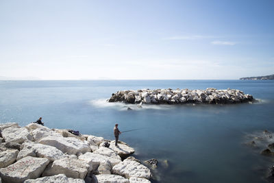 Side view of man standing on rock fishing in sea against sky
