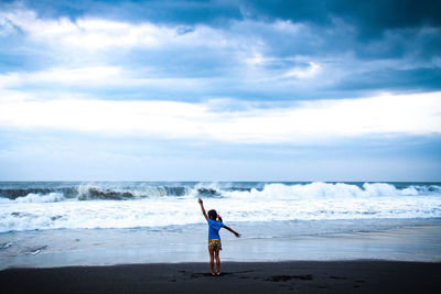 Full length rear view of girl standing on shore at beach against cloudy sky