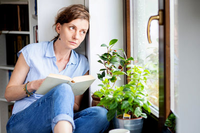 Thoughtful woman with book looking through window at home