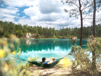 Man relaxing in hammock by lake against cloudy sky