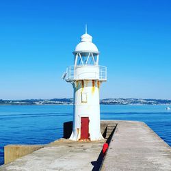 Lighthouse by sea against clear blue sky