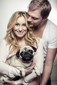 Portrait of happy pregnant woman holding pug dog with man embracing her from behind