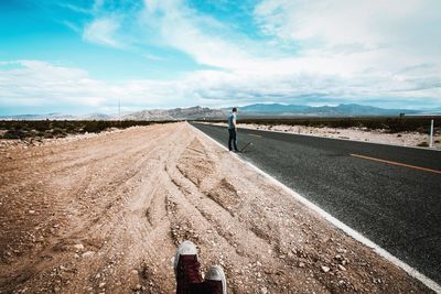 Man with skateboard standing on road against cloudy sky