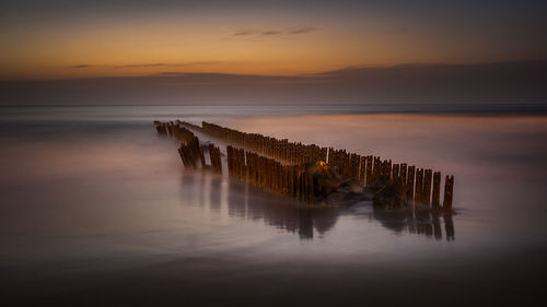 Wooden posts in sea against sky during sunset