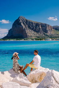 Man sitting on rock by sea against mountains