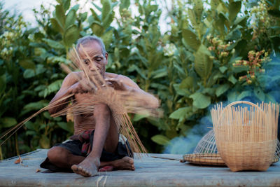 Shirtless senior man making wicker basket while sitting at farm