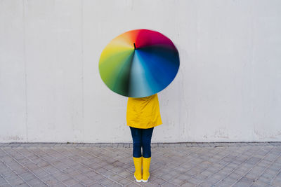 Woman holding colorful umbrella standing in front of gray wall on footpath