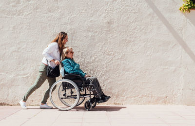 Side view of adult daughter pushing wheelchair with senior mother while walking along city street on sunny day