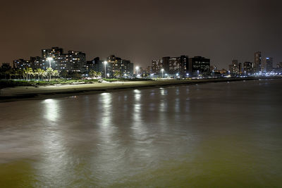 Illuminated buildings by river against sky at night