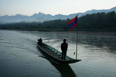 Man standing by flag in boat on lake against mountains