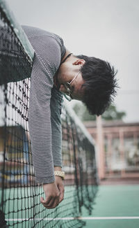 Side view of young man hanging on net against cloudy sky