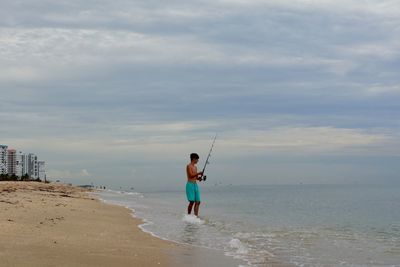 Man fishing while standing on shore at beach against sky