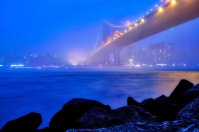 Illuminated suspension bridge over sea against sky at night