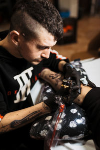 Stylish man with piercing using tattoo machine to make tattoo on leg of crop customer during work in salon