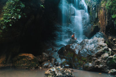 Scenic view of waterfall with man sitting on rock in forest