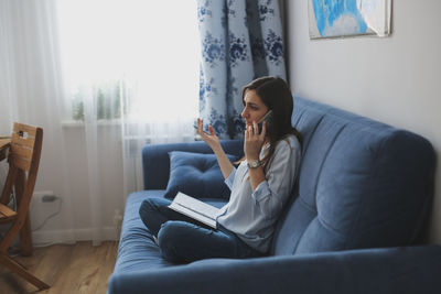 Cute caucasian girl talking on the phone on a blue sofa in a real interior against the window, 