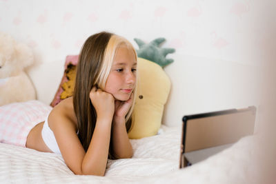 Beautiful teenage girl in pajamas is lying on her bed and watching a video movie on her tablet