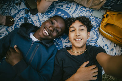 Teenage boy taking selfie with male friend while lying down on picnic blanket