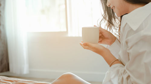 Midsection of smiling woman having coffee while sitting on bed at home