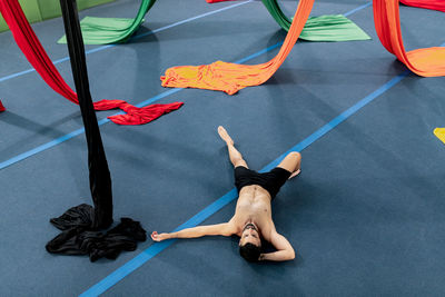 From above shirtless male gymnast lying and relaxing on floor near colorful fabric ribbons during break in aerial dance rehearsal