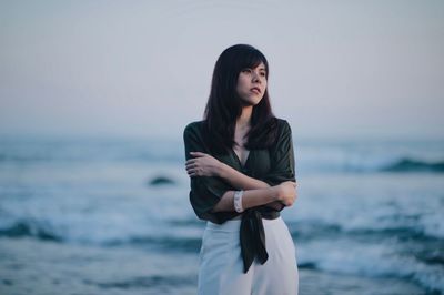 Thoughtful young woman hugging self while looking away at beach against sky