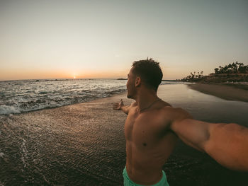 Shirtless man standing at beach against sky during sunset