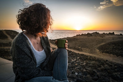 Woman with coffee looking at sea against sky during sunset