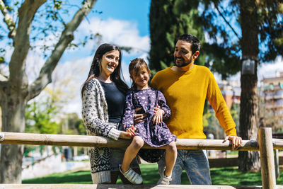 Smiling parents standing by cute daughter sitting on railing in park