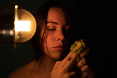 Close-up of young woman holding kiwi by illuminated light bulb