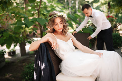 Beautiful bride sitting on chair with groom in background outdoors