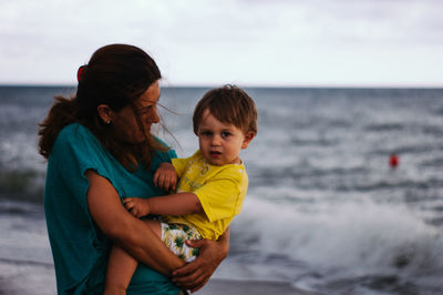 Mother carrying baby boy at seashore against sky